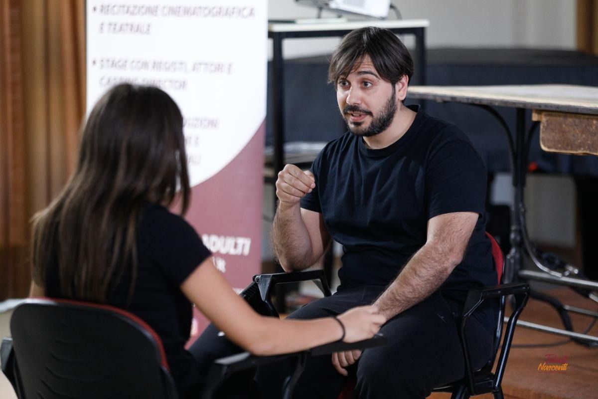 Un uomo gesticola, parlando con una donna. Sono seduti in una stanza, vicino a uno striscione.