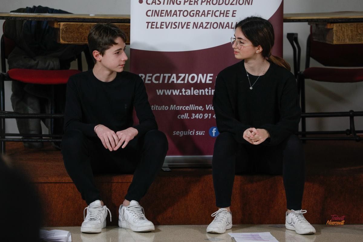 Due persone vestite di nero sono sedute davanti a uno striscione e si guardano.