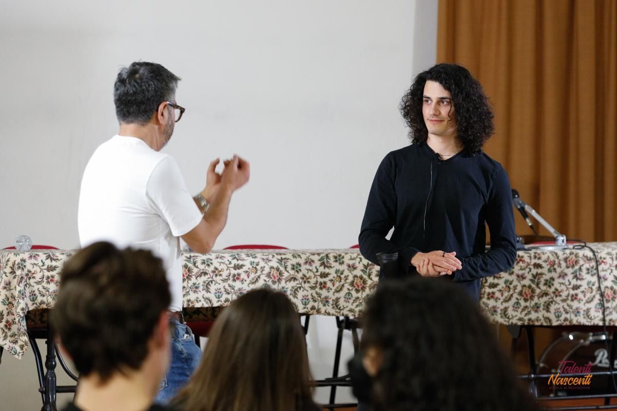 Due persone, una con una camicia bianca e l'altra con una nera, parlano a un tavolo in presenza di un pubblico.