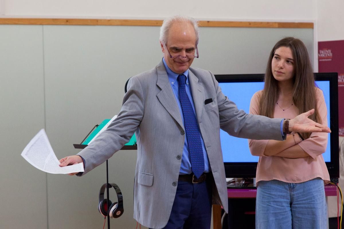Un uomo in giacca e cravatta gesticola con un foglio di carta, una donna è in piedi accanto a lui. All'interno, schermo e leggio sullo sfondo.