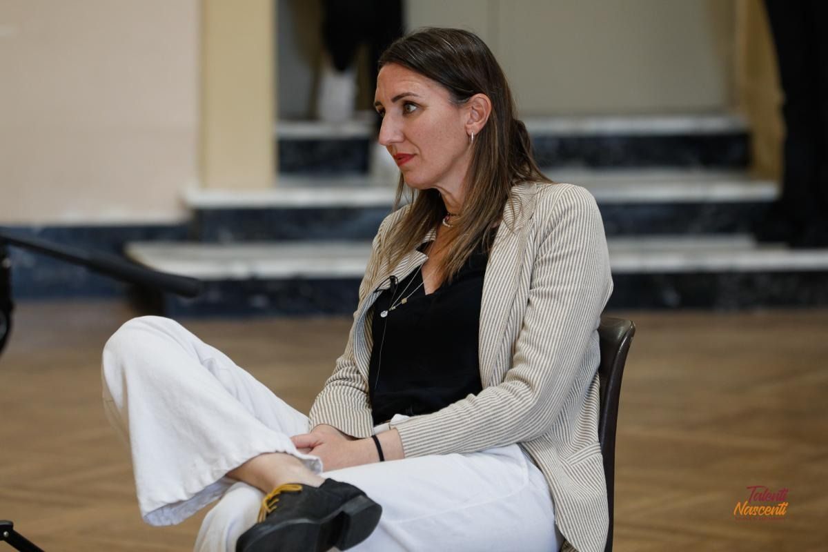 Una donna in pantaloni bianchi e blazer è seduta, con le gambe incrociate, in un interno, e guarda a sinistra.