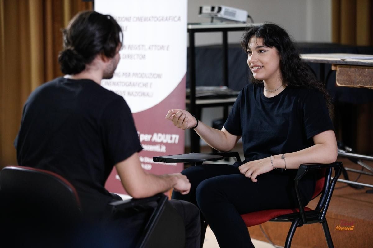 Una donna gesticola mentre parla con un uomo seduto di fronte a lei. Sembra che siano in riunione.