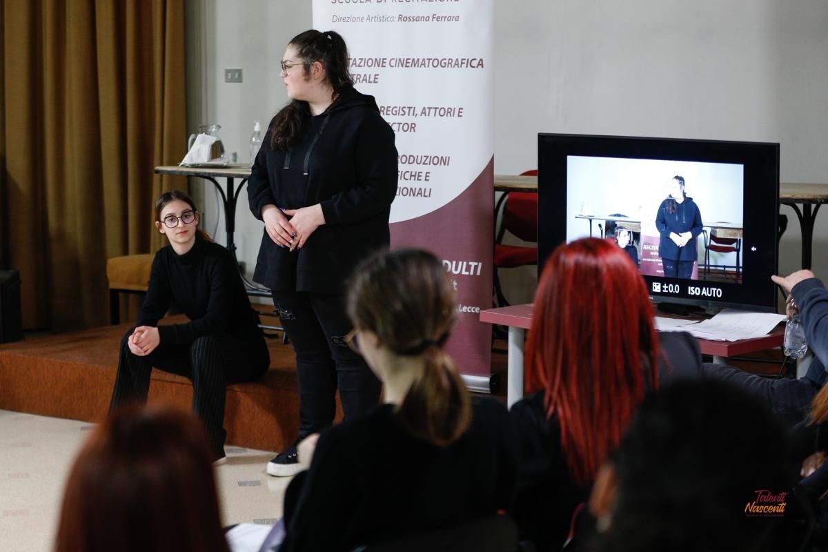 Le persone guardano una presentazione. Due persone sono in piedi su un palco, una delle quali è sullo schermo. Il pubblico è rivolto verso di loro.