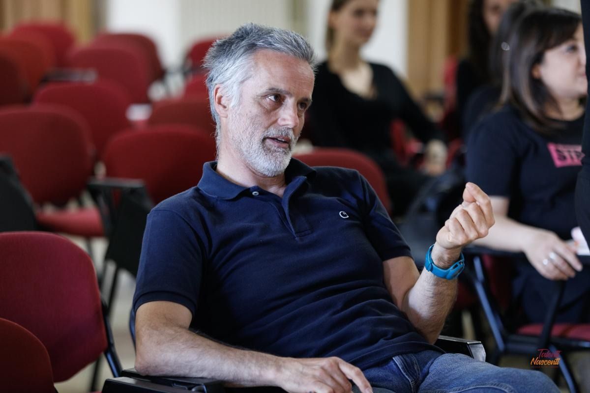 Un uomo con i capelli grigi e una polo blu scuro gesticola mentre è seduto in una stanza con sedie rosse.