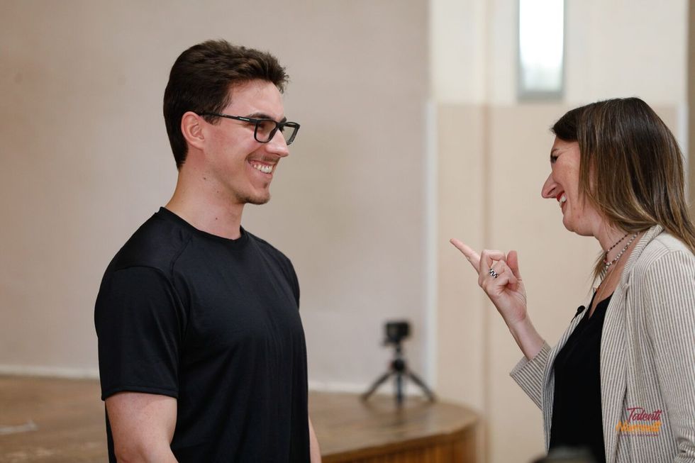 Uomo e donna uno di fronte all'altra, la donna punta il dito. L'uomo sorride, indossa occhiali e camicia nera. Sfondo neutro.