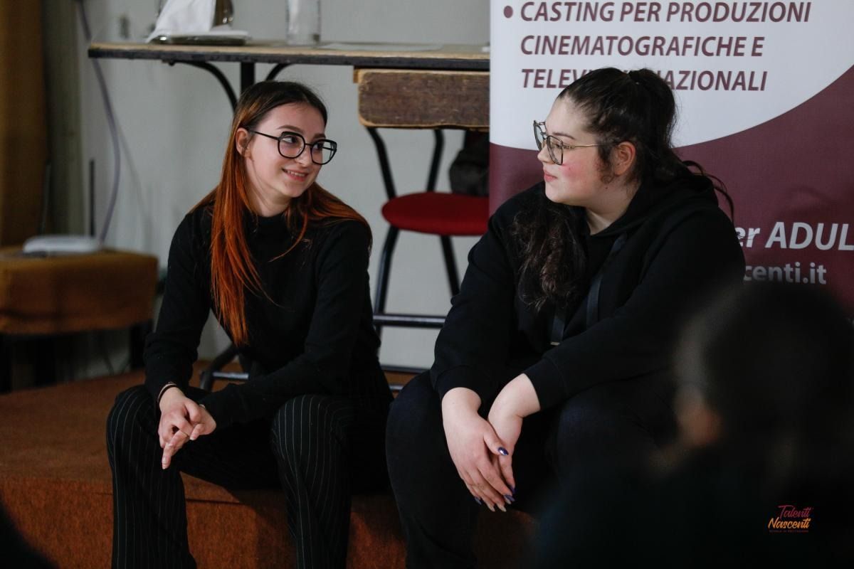 Due persone vestite di nero, sedute e intente a parlare, durante un evento.
