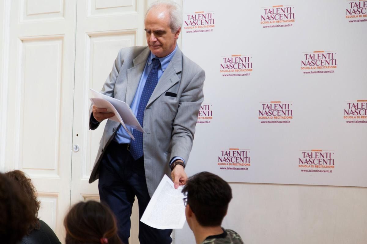 Un uomo in giacca e cravatta porge dei fogli a uno studente davanti a uno sfondo con del testo ripetuto.