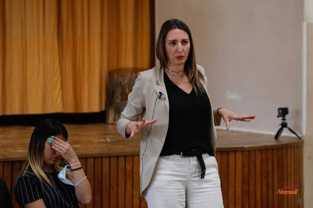 Donna che gesticola durante una presentazione; un'altra donna si copre il volto. Al chiuso, con palco e attrezzatura per la registrazione.