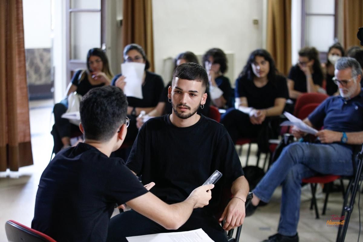 Due uomini in camicia nera sono seduti, uno sta intervistando l'altro. Il pubblico osserva.