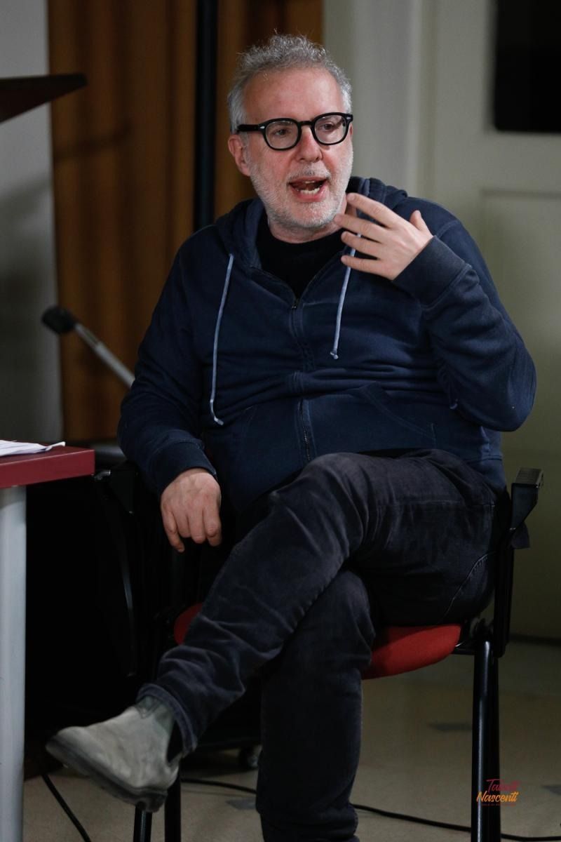 Uomo con occhiali e barba, con felpa blu, che gesticola mentre parla, seduto con le gambe incrociate.