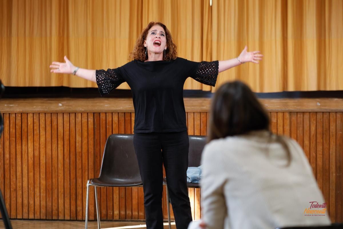 Donna in abito nero con le braccia tese, che si esibisce sul palco con un'altra persona in primo piano.