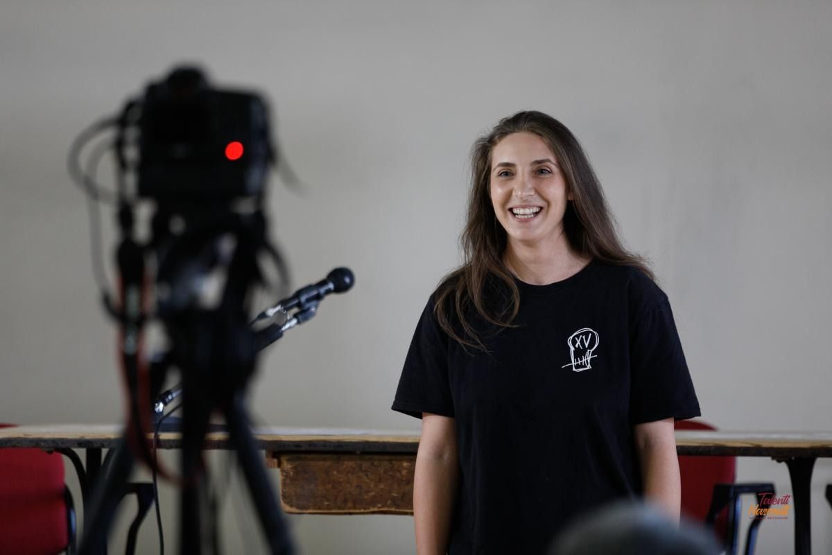 Una donna con una maglietta nera sorride davanti a una telecamera su un treppiede; si trova in una stanza con un tavolo.