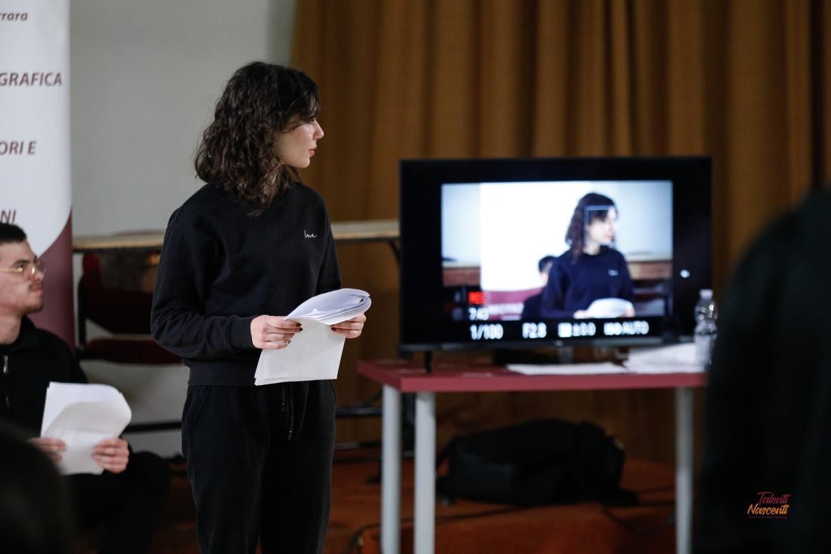 Una donna presenta, con in mano dei documenti, davanti a un monitor che mostra una registrazione in diretta. Un uomo è seduto, con in mano dei documenti, e un semplice tavolo.