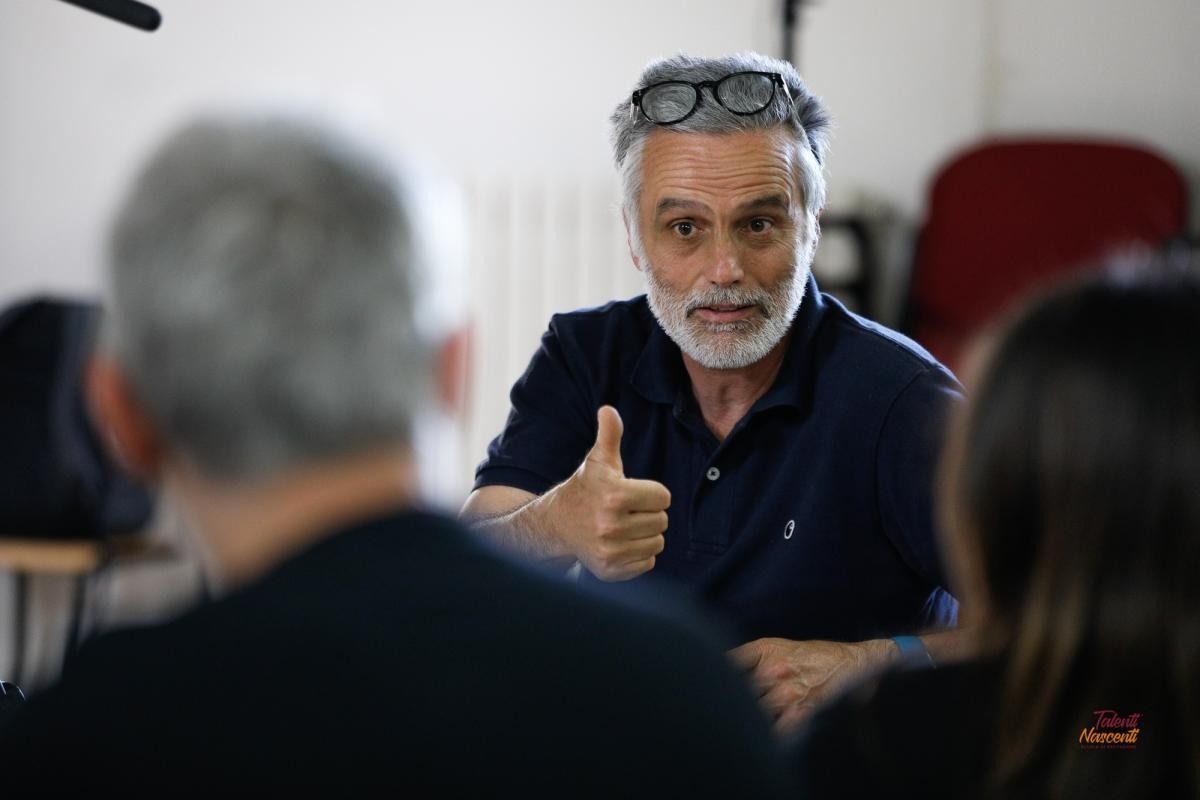 Un uomo con barba e capelli grigi alza il pollice; è seduto, rivolto verso gli altri. Camicia blu, occhiali.
