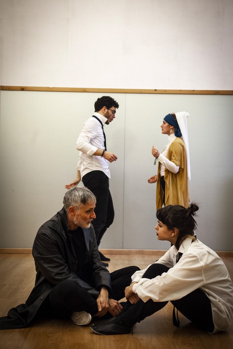 Quattro persone in una stanza, due sedute e due in piedi. Una donna in abiti medievali. Le altre in abiti moderni.