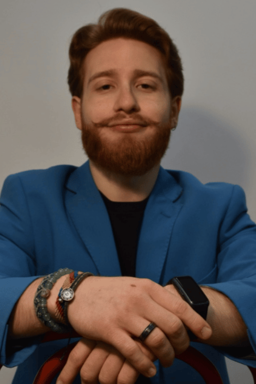 Uomo con barba e capelli rossi, indossa una giacca blu e un orologio, sorride e tiene le braccia incrociate.