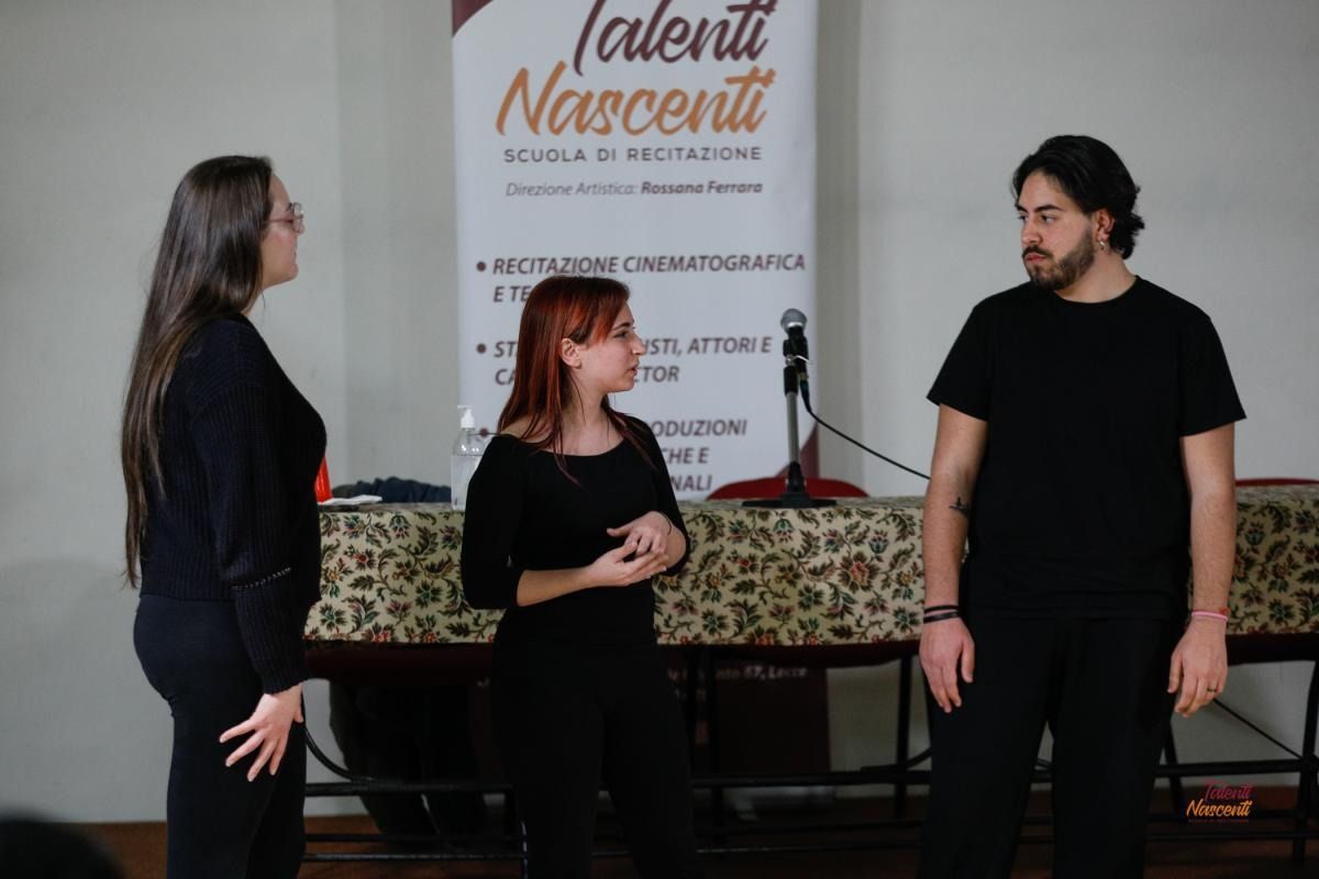 Tre persone davanti a uno striscione. Due parlano e gesticolano; una è in piedi e guarda.