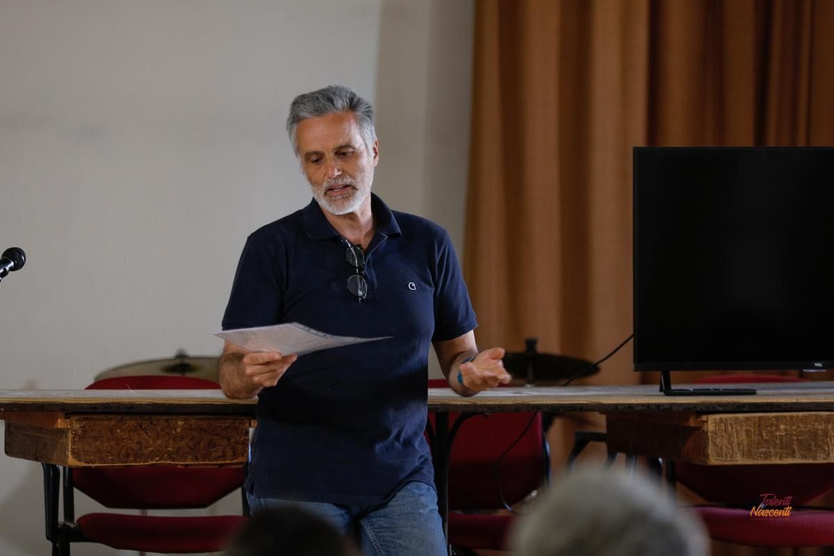Un uomo in camicia blu parla, tenendo in mano un foglio; è dietro un tavolo, accanto a una TV, in un ambiente interno.