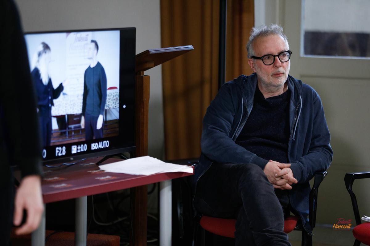 Un uomo con gli occhiali guarda un monitor che mostra una scena in bianco e nero; è seduto a un tavolo con dei documenti.