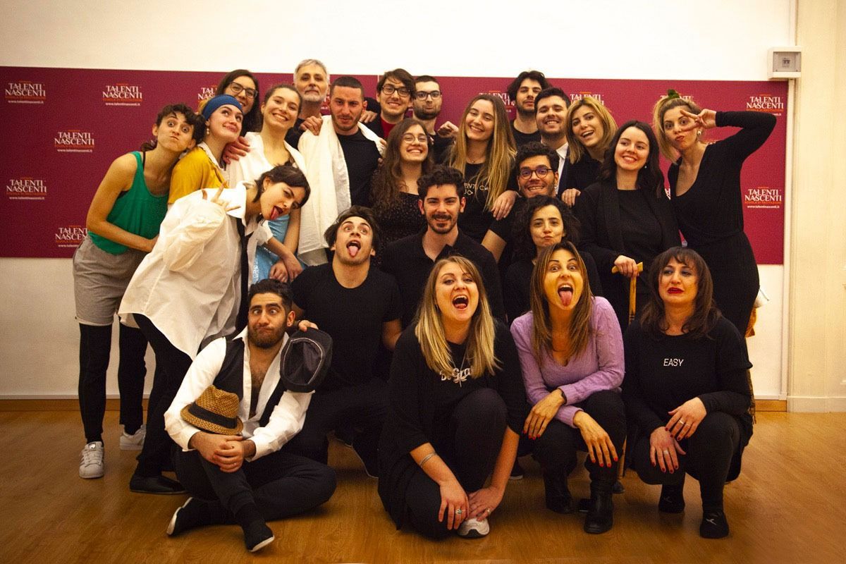 Un gruppo di persone in posa, alcune con la lingua fuori, davanti a un muro color marrone con dei loghi.