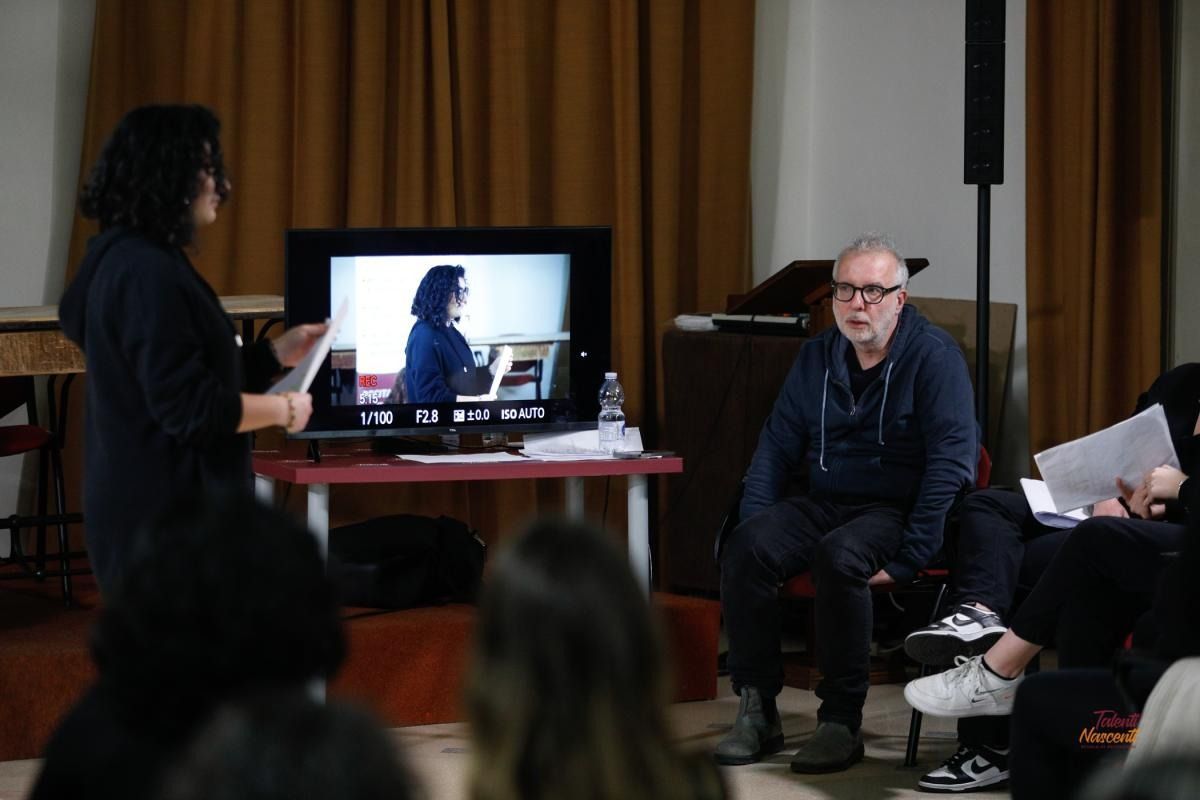 Una donna presenta un video a un gruppo in una stanza, mentre uno schermo mostra un video. Un uomo guarda seduto.