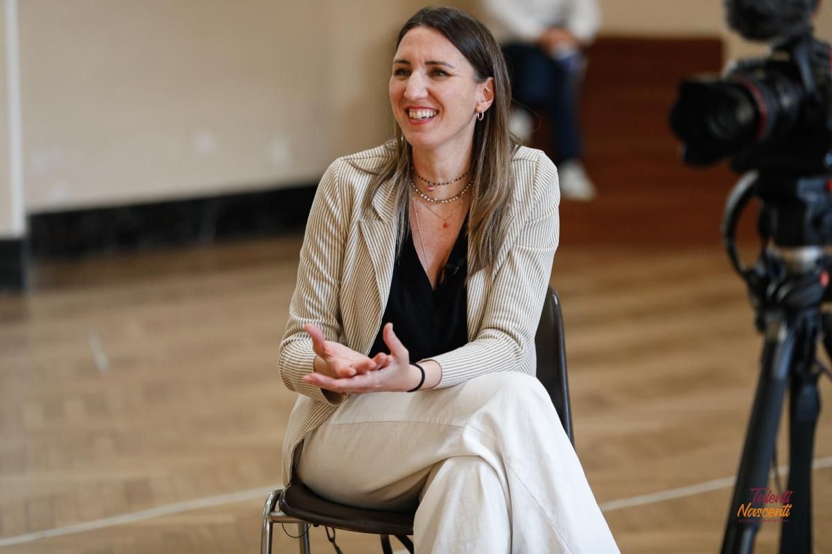 Una donna con una giacca leggera e pantaloni sorride, seduta su una sedia, gesticolando con le mani durante un'intervista.