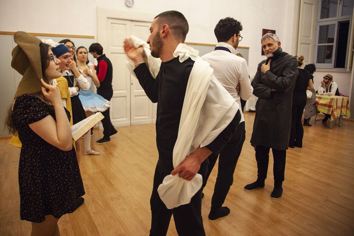 Persone in costume sono in una stanza. Un uomo fa un gesto verso una donna che indossa un indumento marrone con cappuccio. Altri guardano.