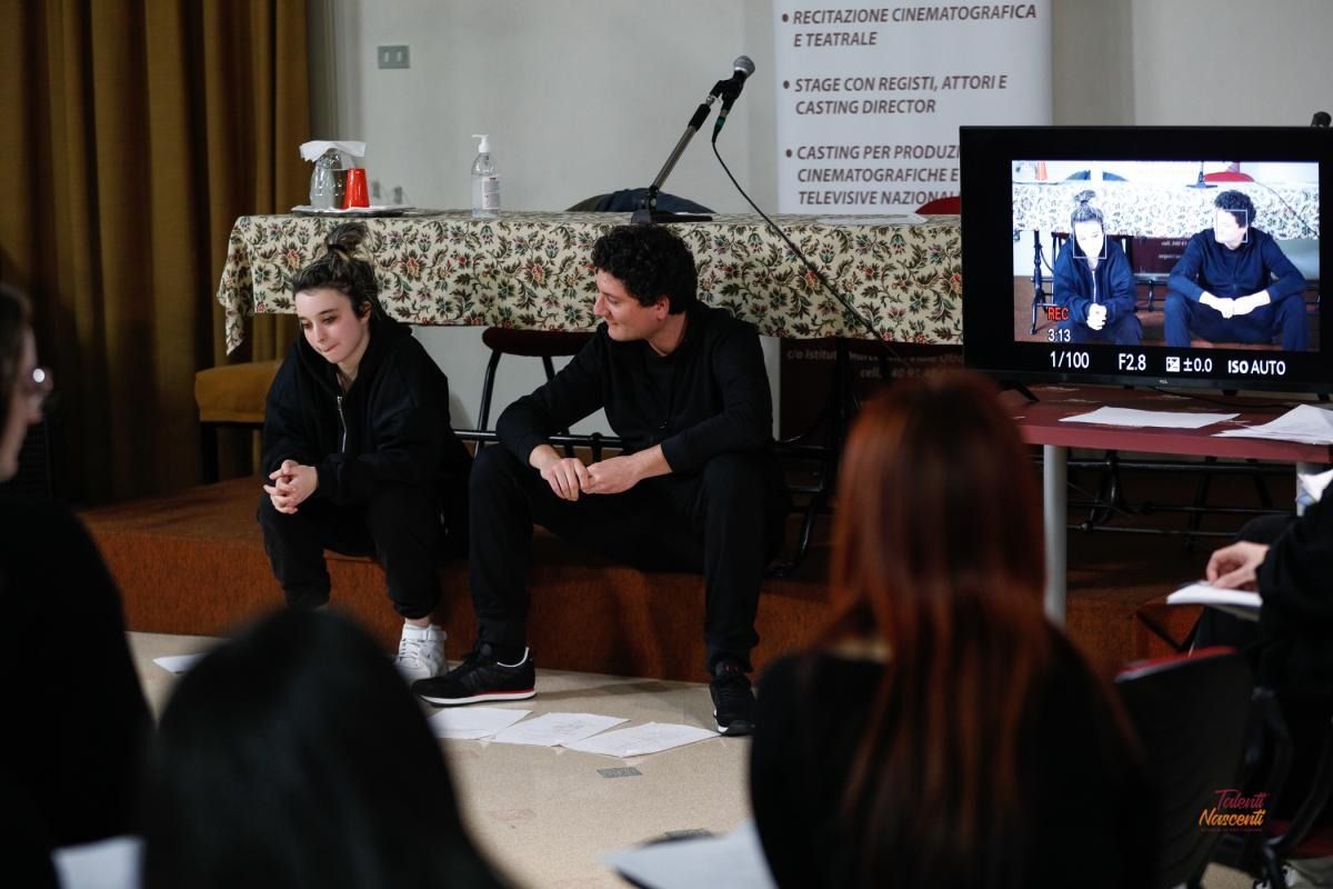 Due persone in abiti neri, sedute durante una presentazione, inquadrate da un monitor. Il pubblico è visibile in primo piano.