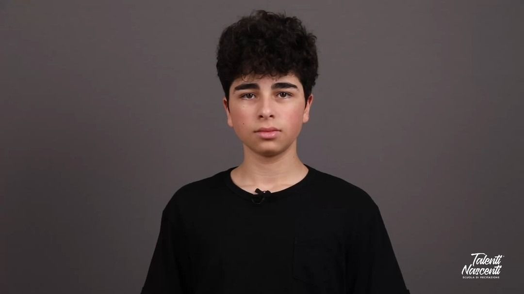 Adolescente con capelli scuri e ricci, maglietta nera su sfondo grigio, che guarda la telecamera.