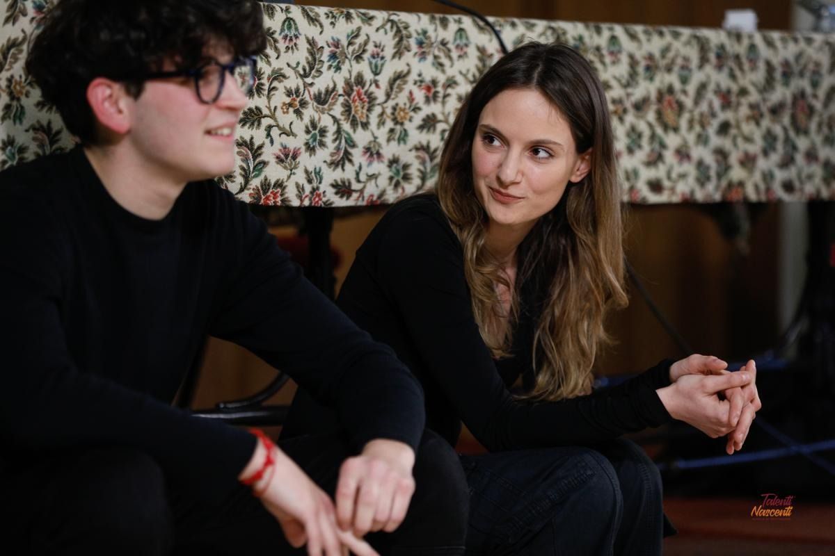 Due persone sedute, che parlano. Una indossa gli occhiali e sorride; l'altra ha i capelli lunghi e guarda, con le braccia appoggiate. In un interno, su uno sfondo a motivi.