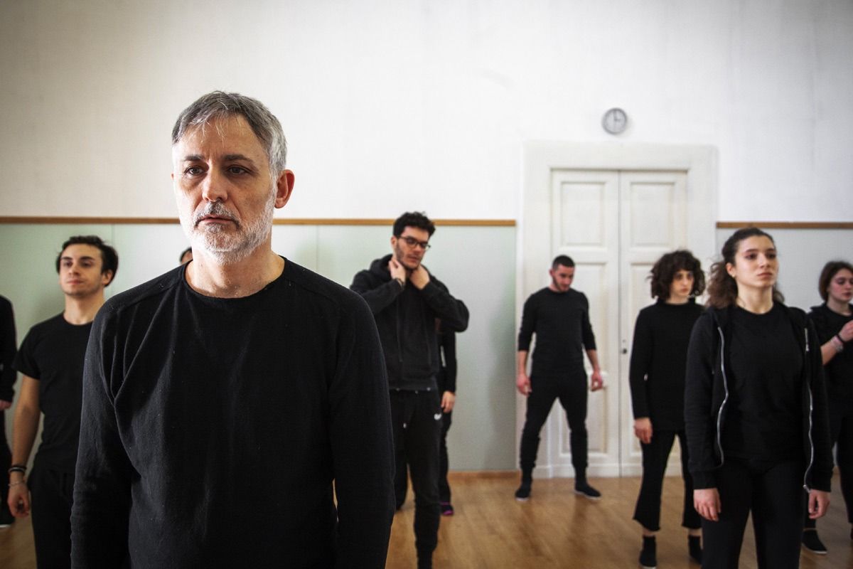 Un uomo e un gruppo di persone in abiti neri, in un interno. L'uomo in primo piano guarda avanti con un'espressione seria.