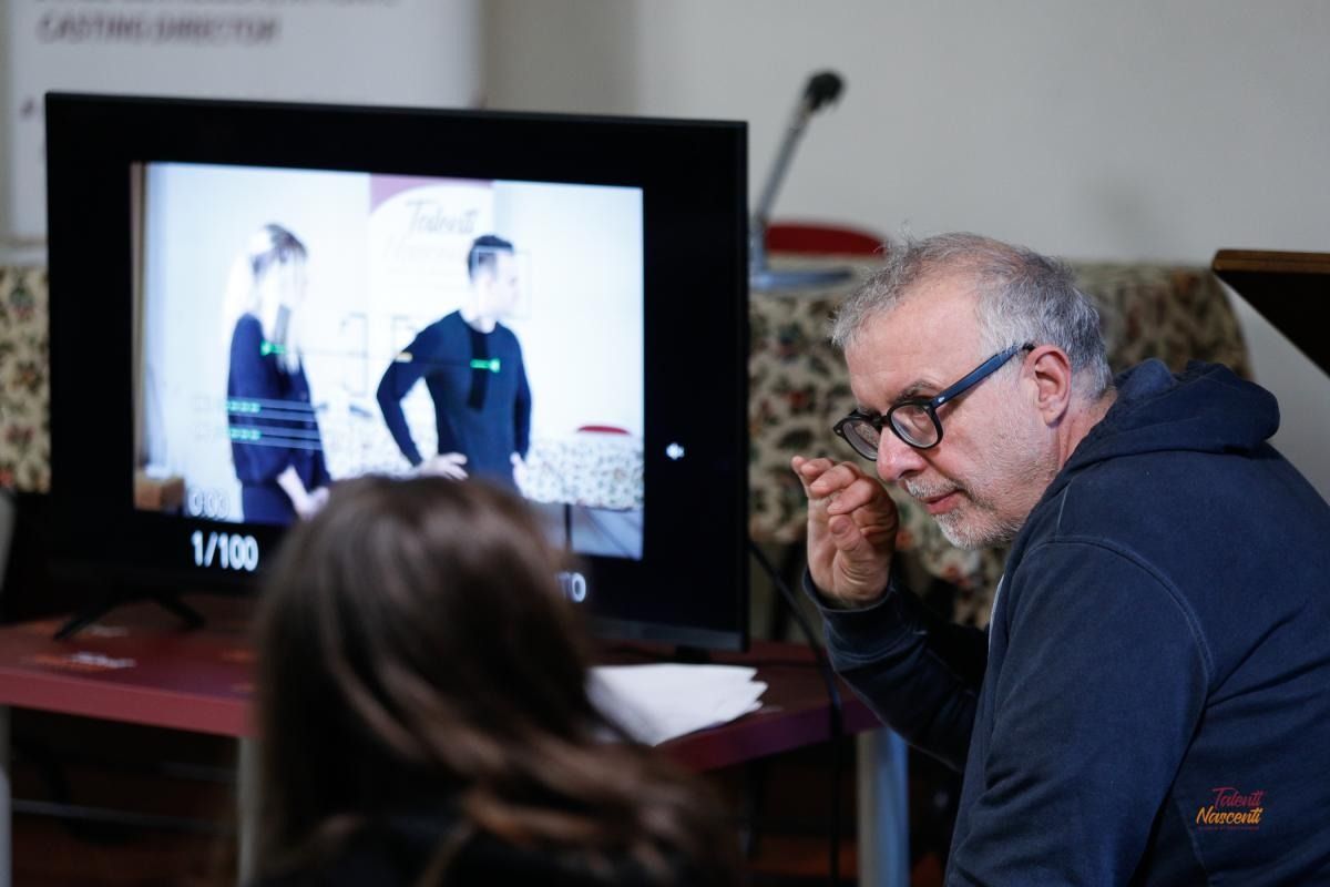 Un uomo con gli occhiali guarda uno schermo su cui sono visualizzate due persone; in primo piano c'è la schiena di una donna.