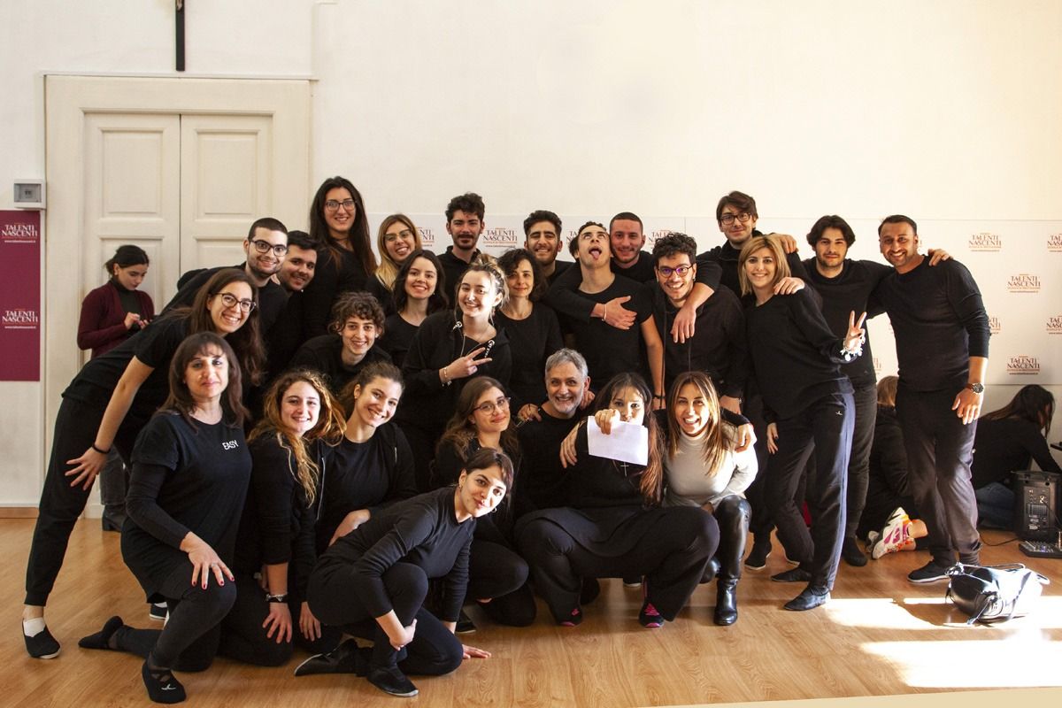 Gruppo di persone in abiti neri, sorridenti. All'interno di un edificio, probabilmente durante una prova.