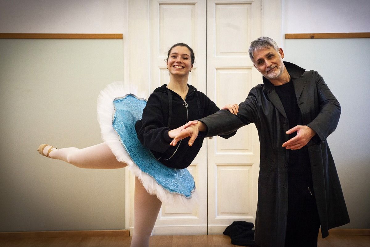 Una ballerina in tutù blu è in equilibrio, tenuta da un uomo con un cappotto, in una sala da danza.
