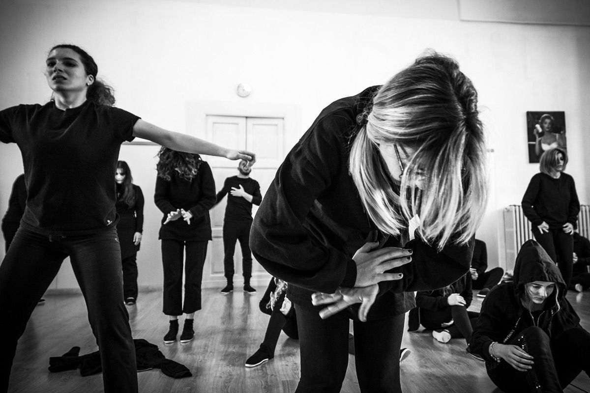 Gruppo di persone in abiti neri in uno studio, alcune si inchinano, altre tengono le braccia tese.