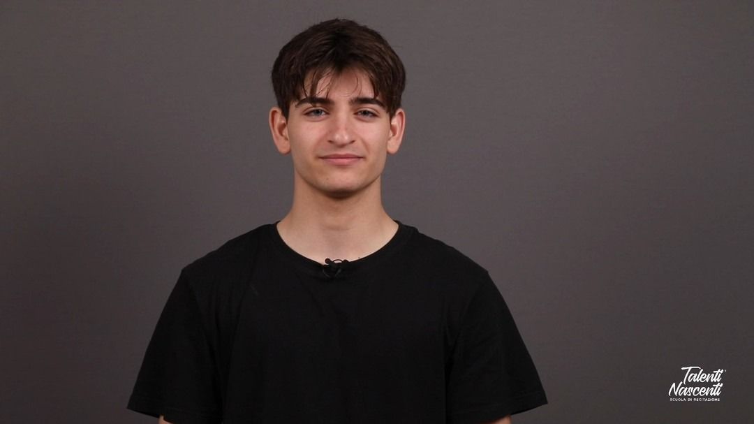 Uomo con camicia nera che sorride alla telecamera su uno sfondo grigio.