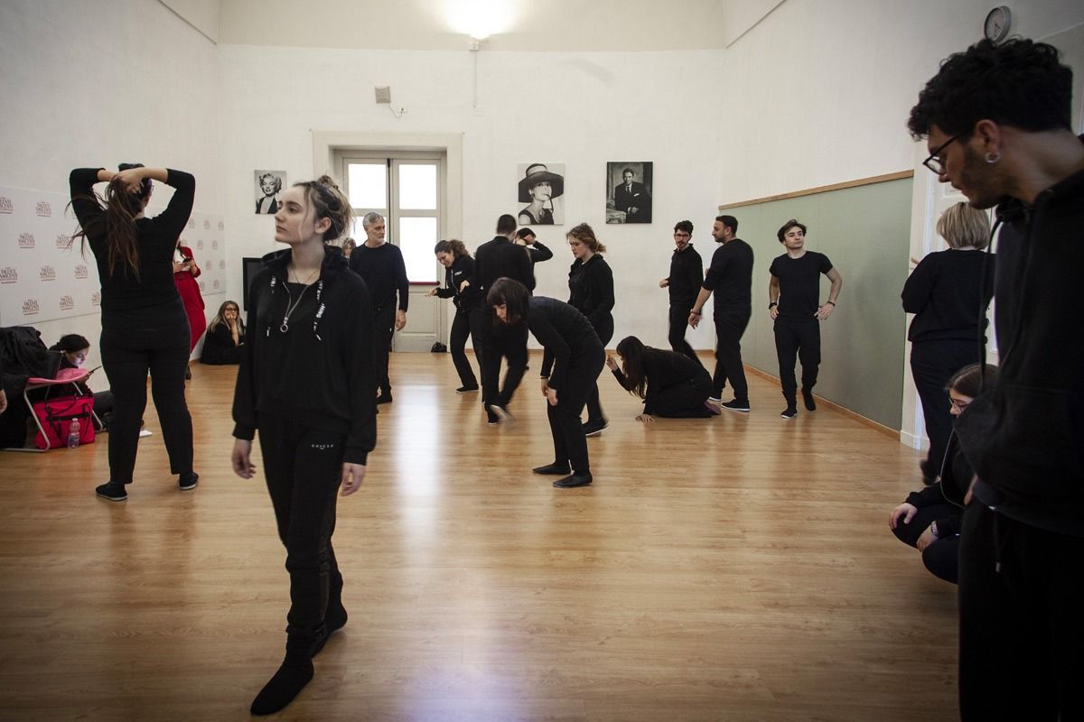 Persone vestite di nero provano uno spettacolo in una grande sala con pavimenti in legno.