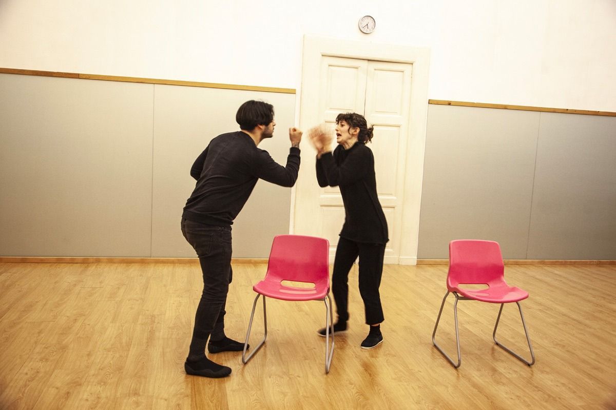 Due persone vestite di nero mimano l'interazione con una palla in una stanza con sedie rosa.