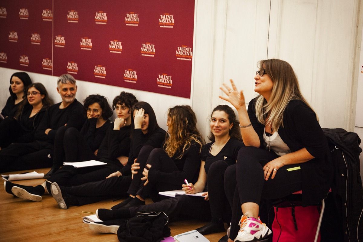 Un gruppo di persone è seduto sul pavimento, ascoltando una donna che gesticola. Sullo sfondo, un muro rosso.