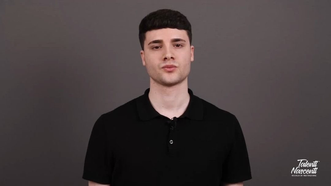 Un uomo con una camicia nera su sfondo grigio parla direttamente alla telecamera.