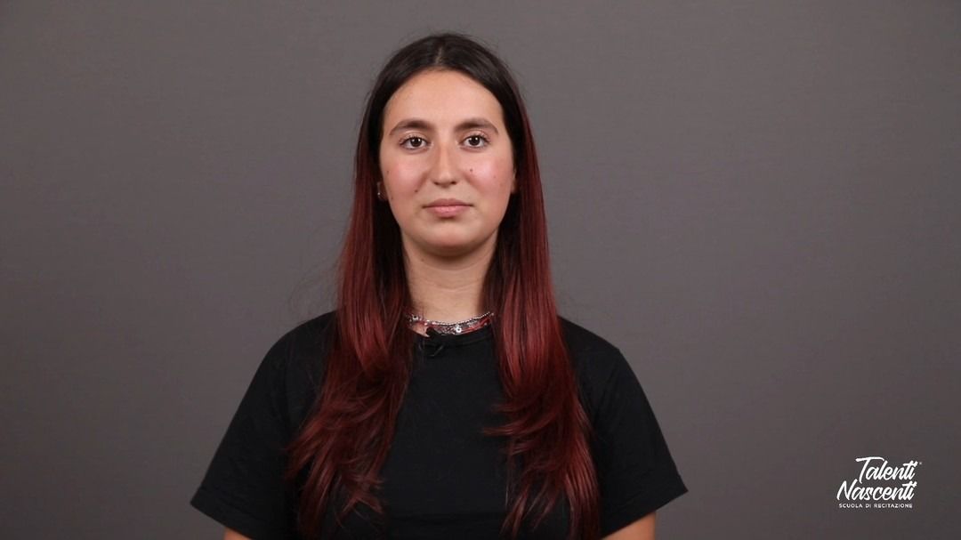 Donna con lunghi capelli scuri con riflessi rossi, che indossa una camicia nera, guarda la telecamera. Sfondo grigio.