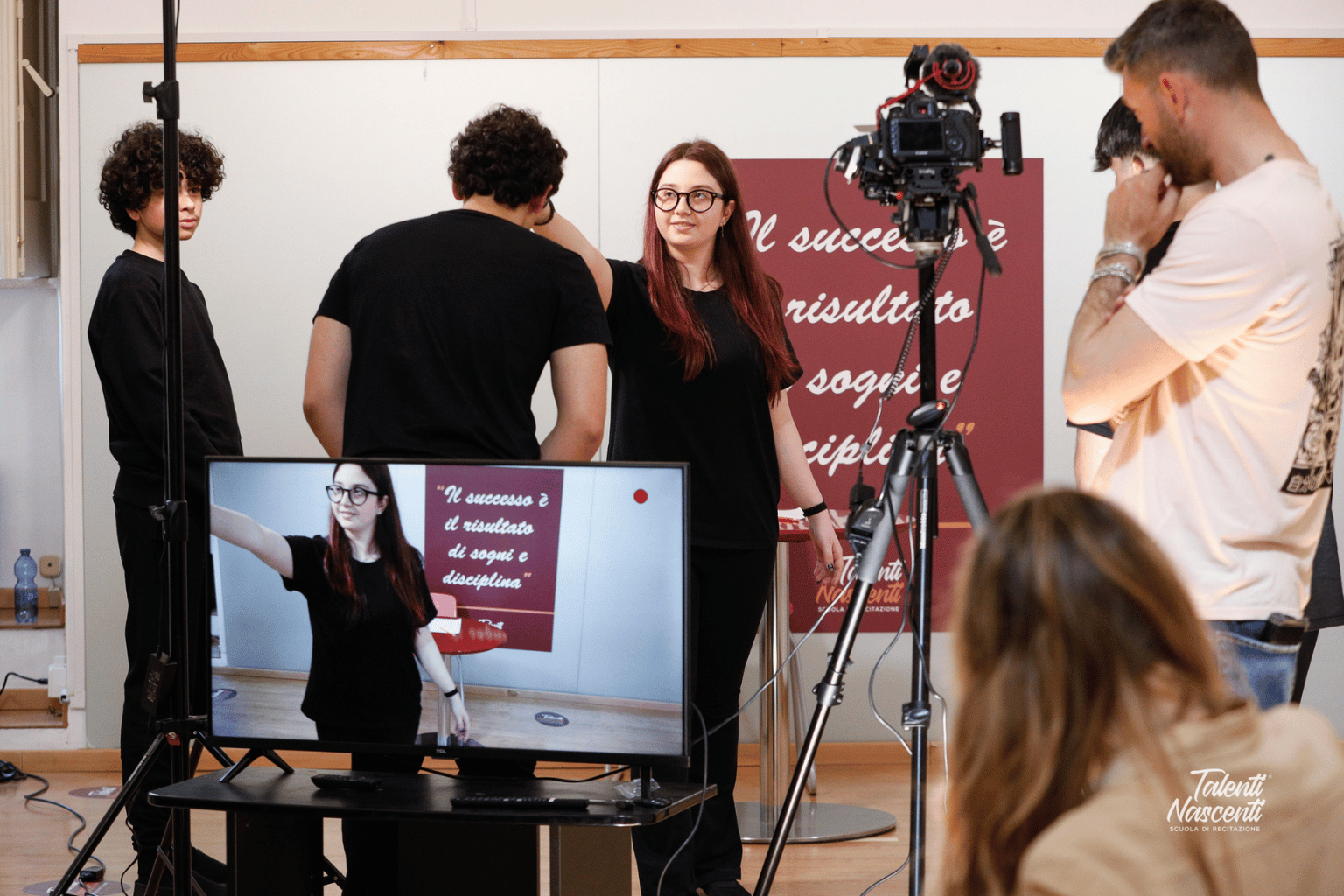 Donna che filma con una troupe, monitor che mostra la sua immagine. Cartello rosso con testo. Ambientazione in studio.