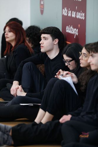 Persone vestite di nero siedono sul pavimento, alcune guardano avanti, altre scrivono. Sullo sfondo è visibile un cartello rosso.