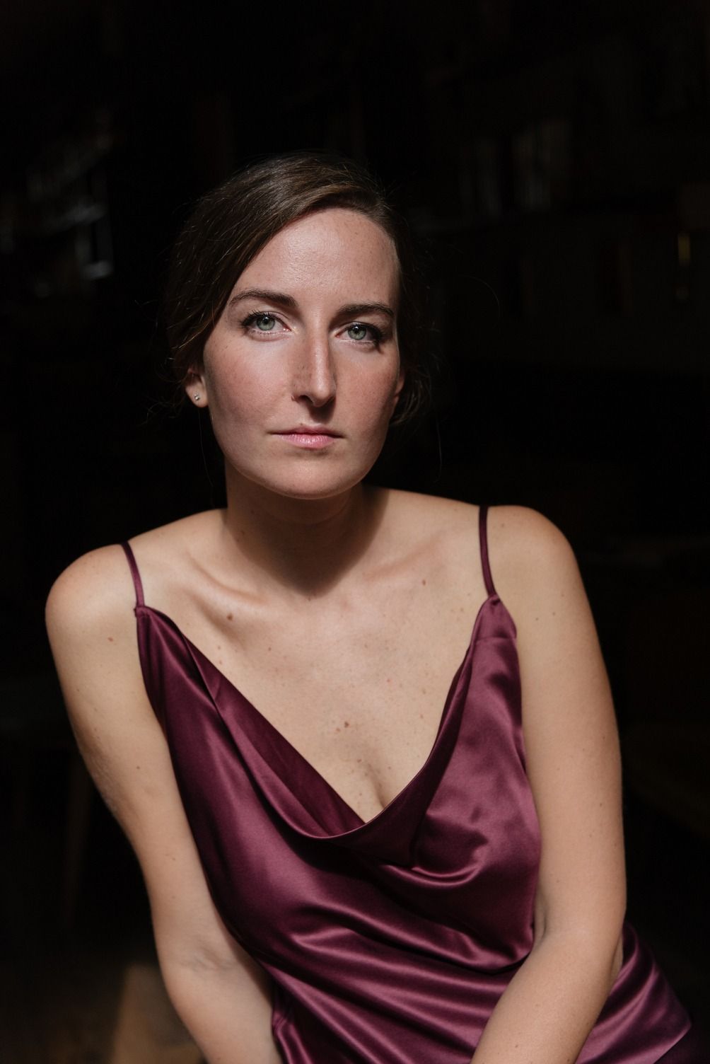 Donna con abito di raso color bordeaux, sguardo serio in avanti, sfondo scuro.