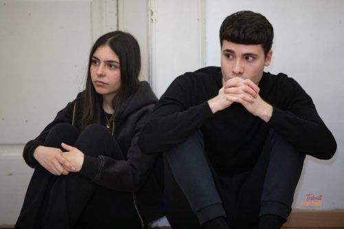 Due persone sono sedute vicine e guardano lontano. Una donna con una felpa nera, un uomo con una maglietta nera, in un interno.