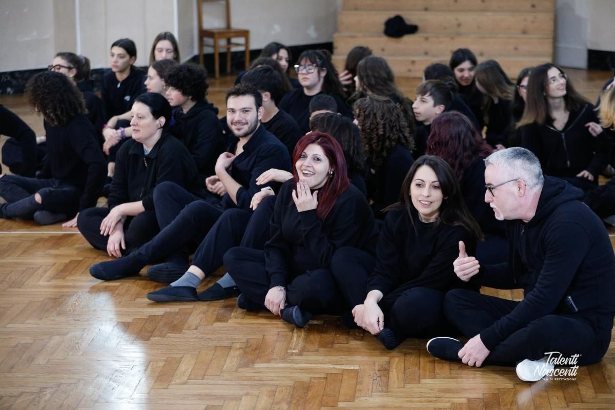 Gruppo di persone vestite di nero sedute su un pavimento di legno, alcune delle quali sorridono.