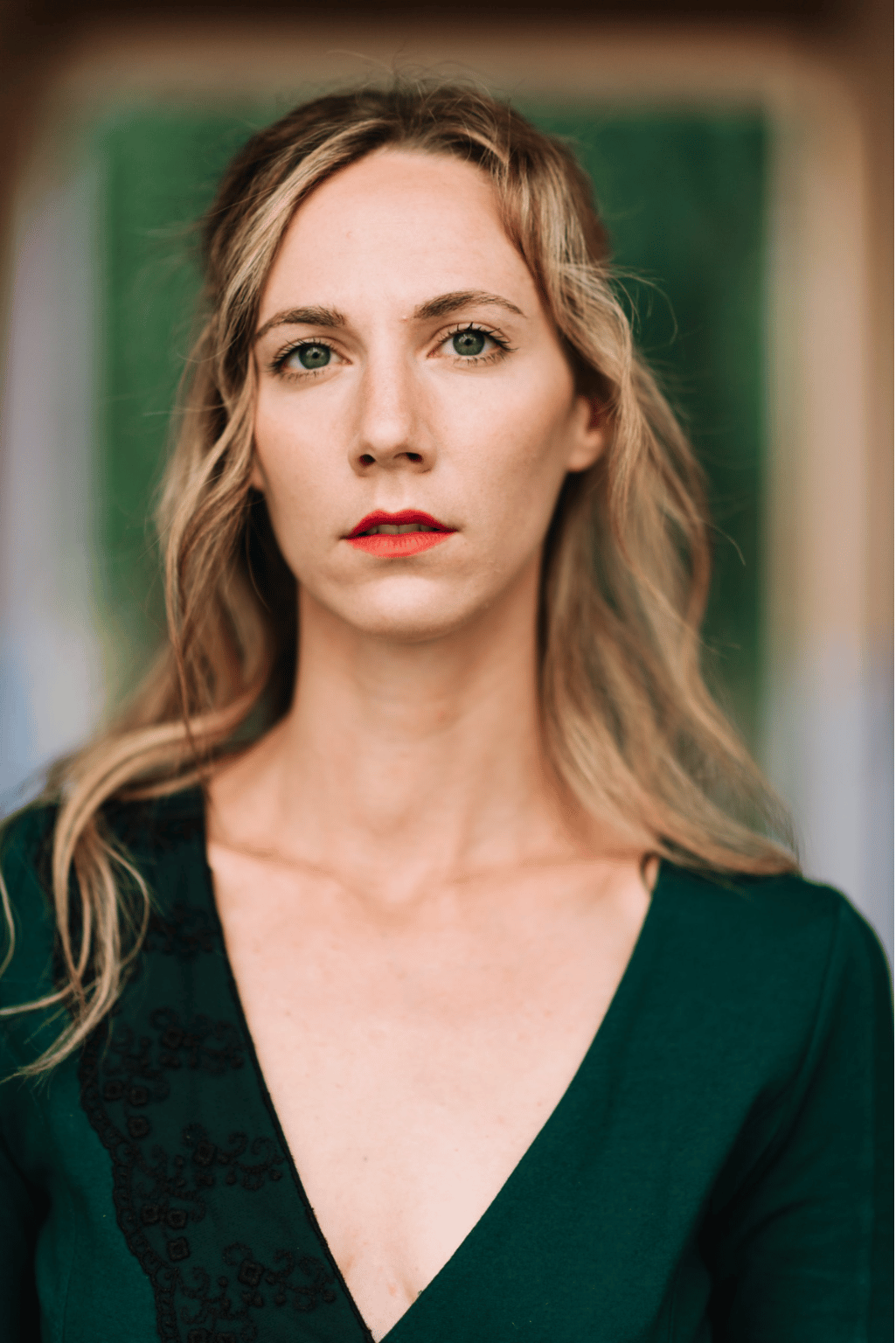Donna con capelli biondi e rossetto rosso, che indossa un abito verde scuro e guarda direttamente l'osservatore.