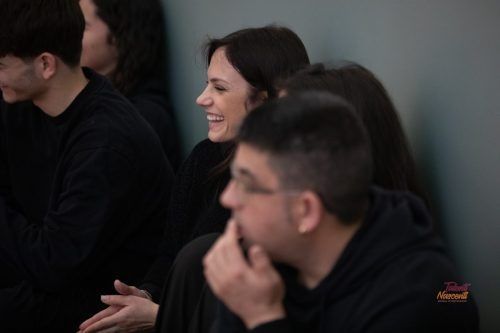 Una donna sorride, seduta tra altre persone vestite di nero, in un interno.