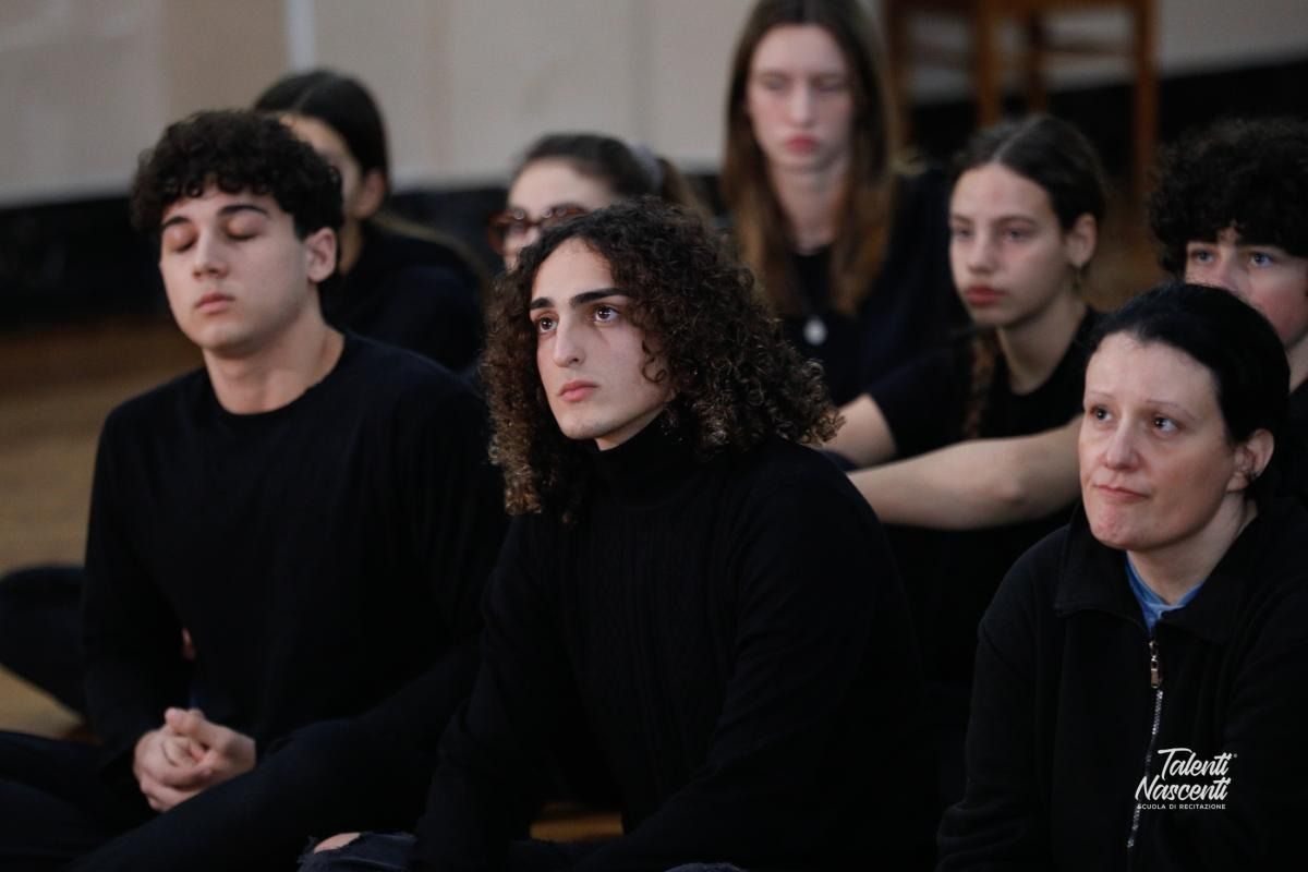 Un gruppo di persone sedute, alcune con gli occhi chiusi. Per lo più vestite di nero, in una stanza.