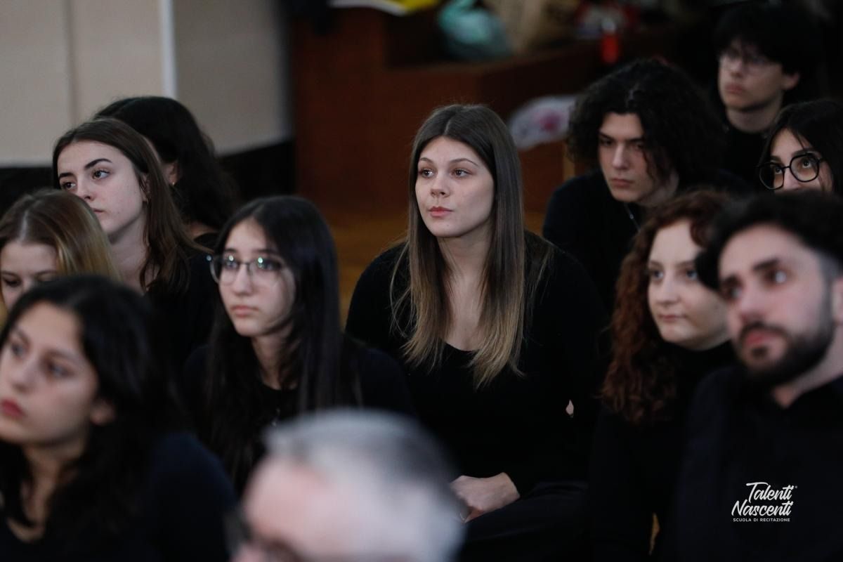 Pubblico seduto, molti in abiti neri, che osserva attentamente qualcosa. All'interno, probabilmente una presentazione.